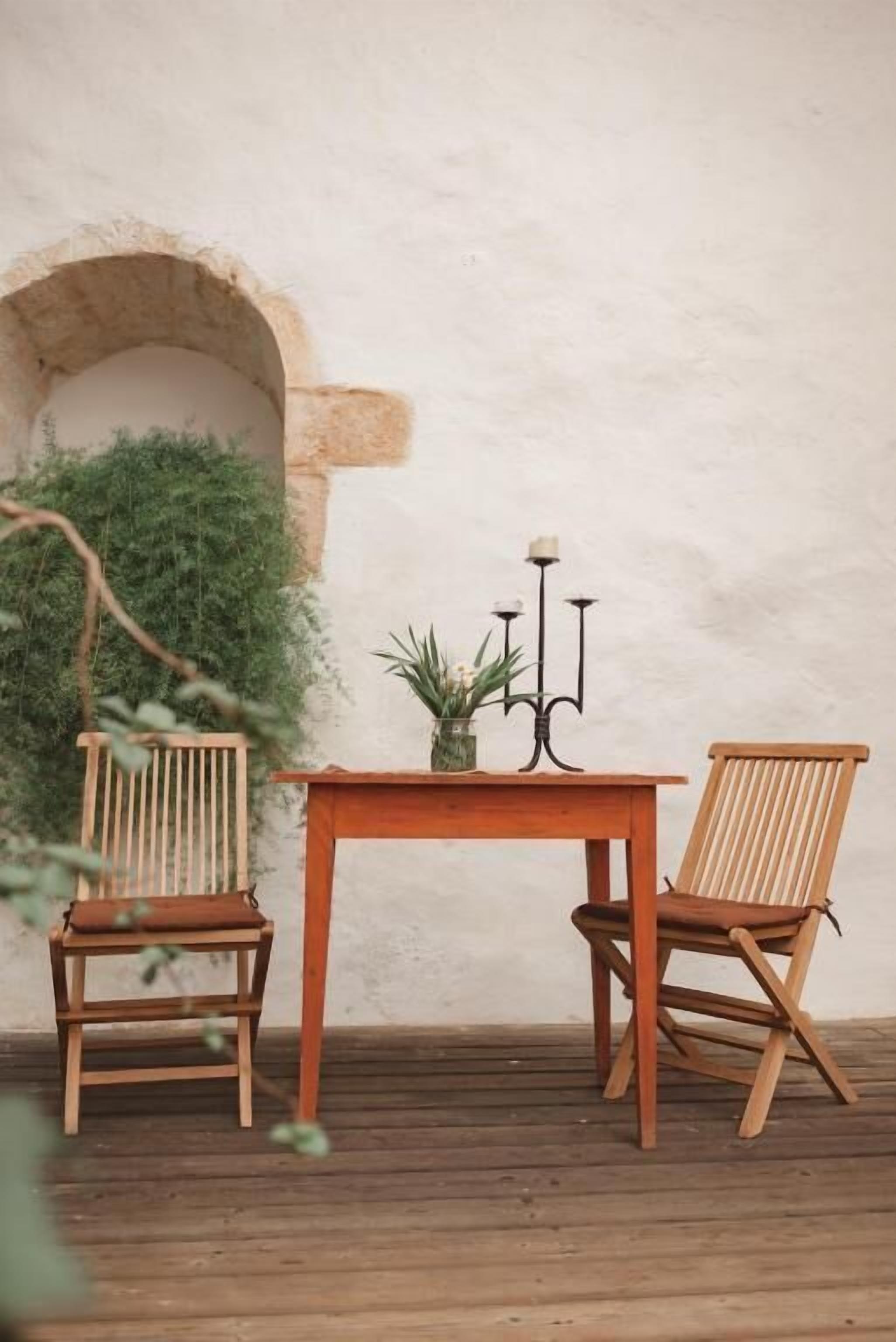 Una mesa de madera con dos sillas, un candelabro y una planta en maceta sobre una terraza de madera frente a una pared blanca con un arco.