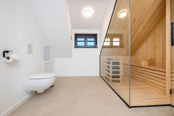 Un baño moderno con inodoro, pared blanca, ventana y zona de sauna de madera.