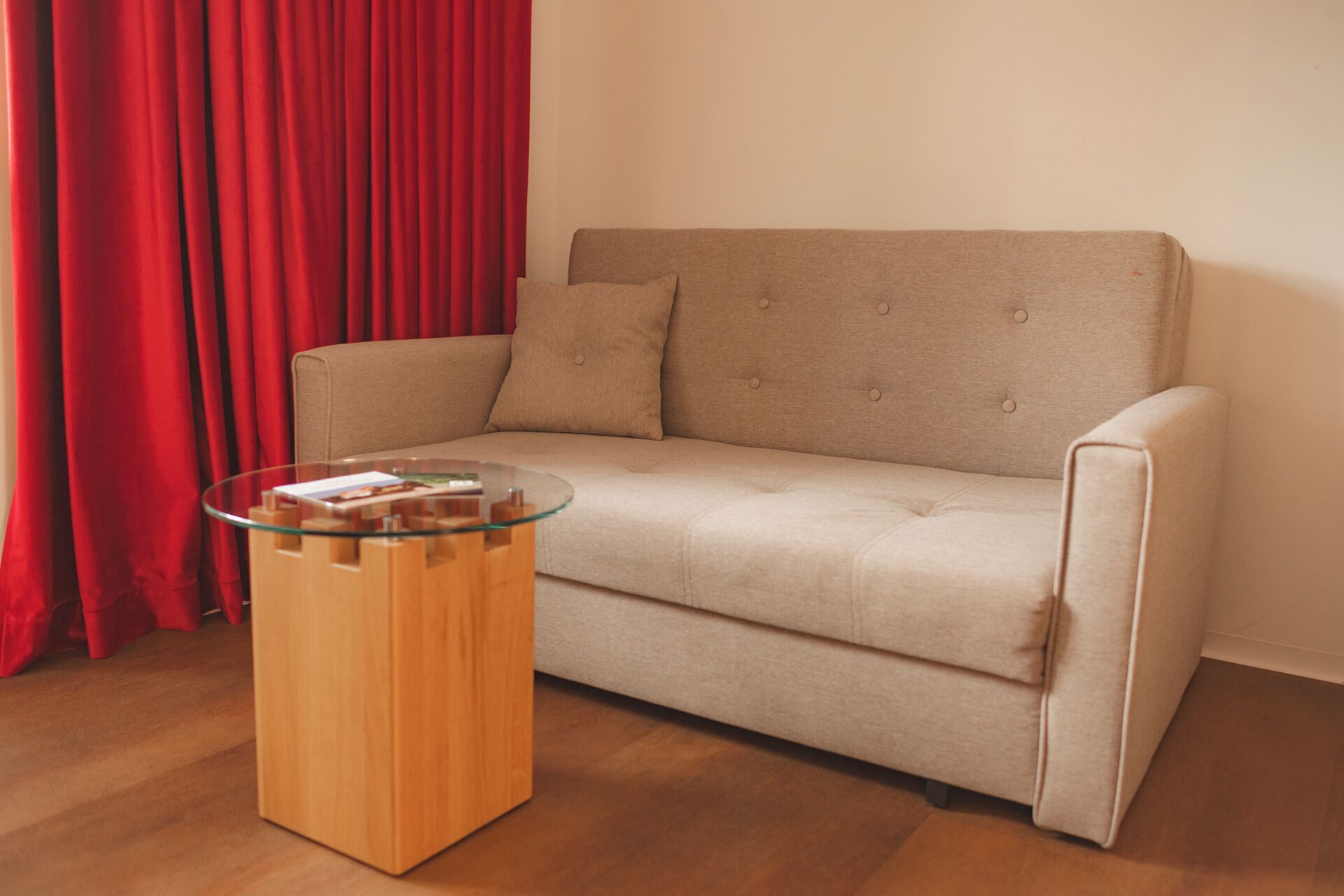 A beige sofa with two cushions, a glass coffee table with books, and red curtains.