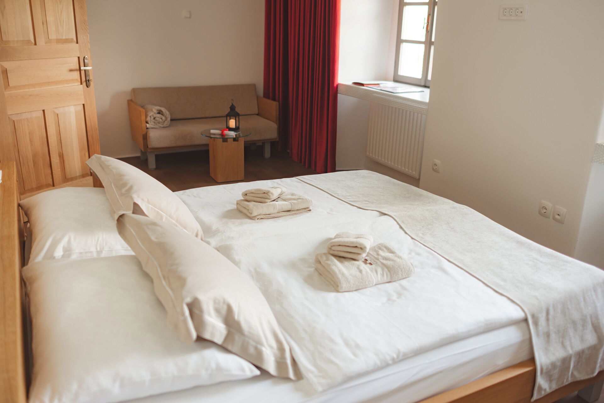 A neatly made bed with white linens and towels, a wooden wardrobe, a sofa, and a window with red curtains.