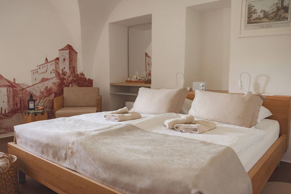 Una cama doble con sábanas y almohadas blancas, un cabecero de madera, una mesita de noche con lámpara y un mural en la pared con un castillo.
