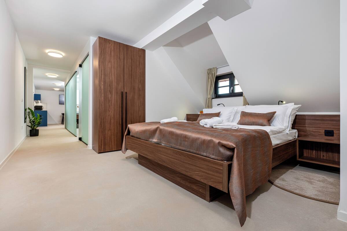 Un dormitorio moderno con una cama de madera, una mesita de noche y una lámpara de noche. Hay una ventana con cortinas, una lámpara en el techo y una planta en la esquina.