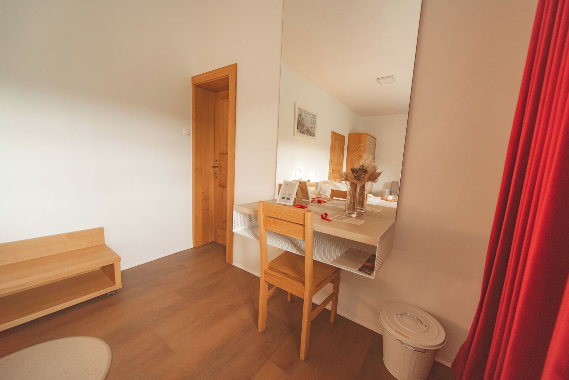 A compact hotel room with a wooden desk, a chair, a small bedside table, a mirror, a red curtain, and a trash bin.