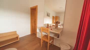 A compact hotel room with a wooden desk, a chair, a small bedside table, a mirror, a red curtain, and a trash bin.