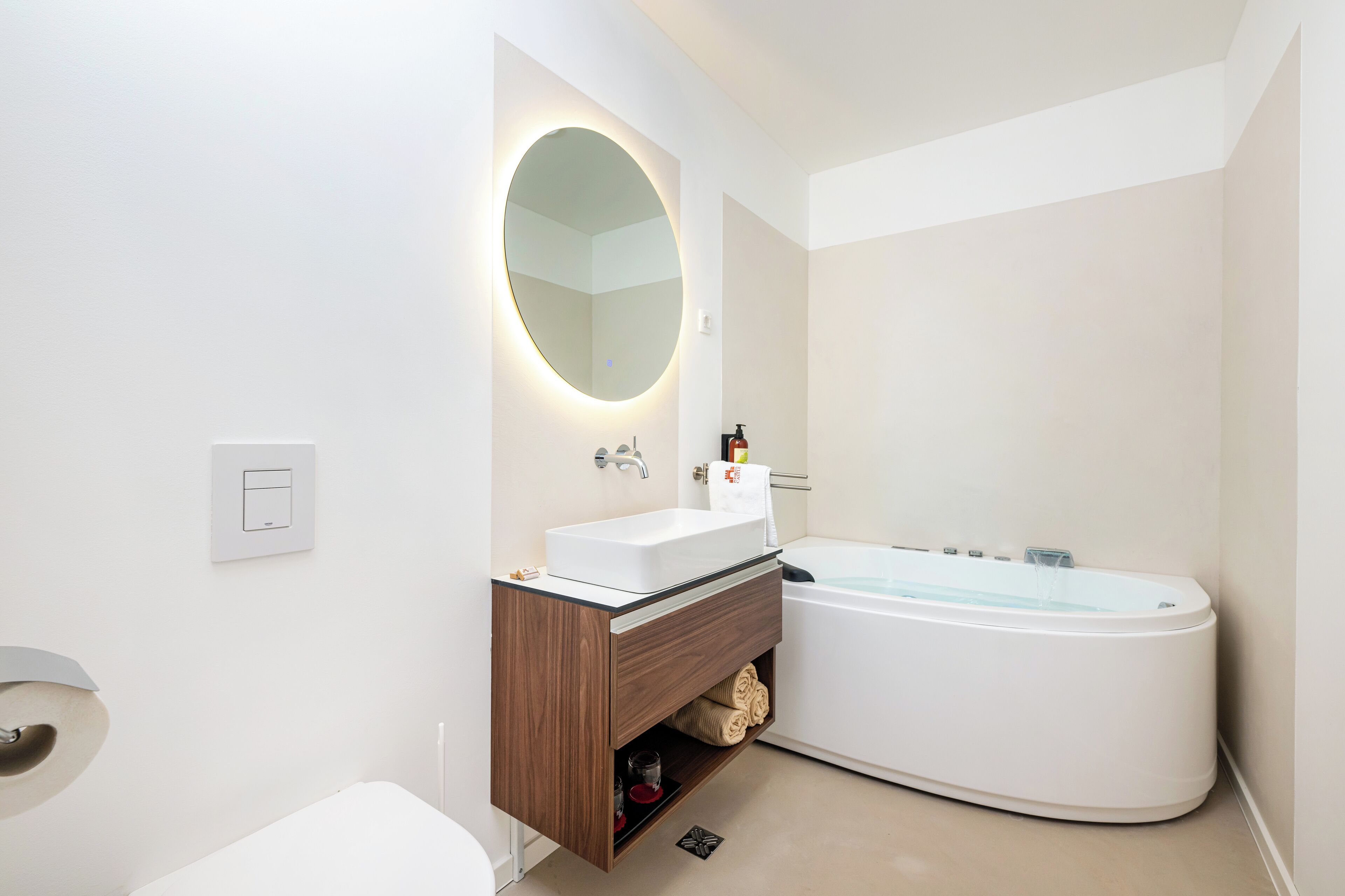 Un baño moderno con un lavamanos blanco, un espejo redondo, una bañera empotrada y un mueble de tocador de madera.