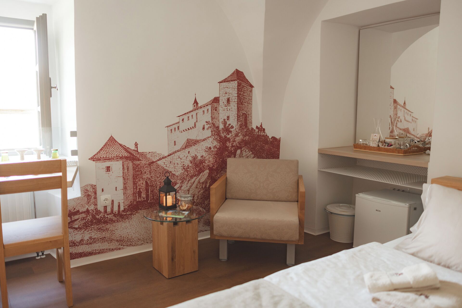 A bedroom with a bed, a chair, a small table with a candle, a mirror, and a wall with a red mural.