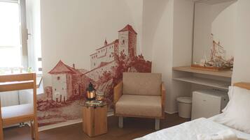 A bedroom with a bed, a chair, a small table with a candle, a mirror, and a wall with a red mural.
