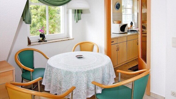 A dining area with a round table, four chairs, a window with a view, and a kitchen visible through an open door.
