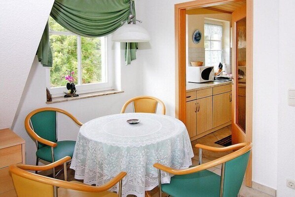 A dining area with a round table, four chairs, a window with a view, and a kitchen visible through an open door. - Charmante Ferienwohnung auf Rügen, Altenkirchen (Altenkirchen)