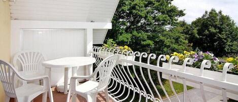 A balcony with white chairs and a table, a white railing, and a view of trees and flowers.