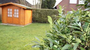 A wooden shed with a door and windows, a well-maintained lawn, and a hedge in the background. - Doppelhaushälfte Friesenglück mit Sauna (Dornumersiel)