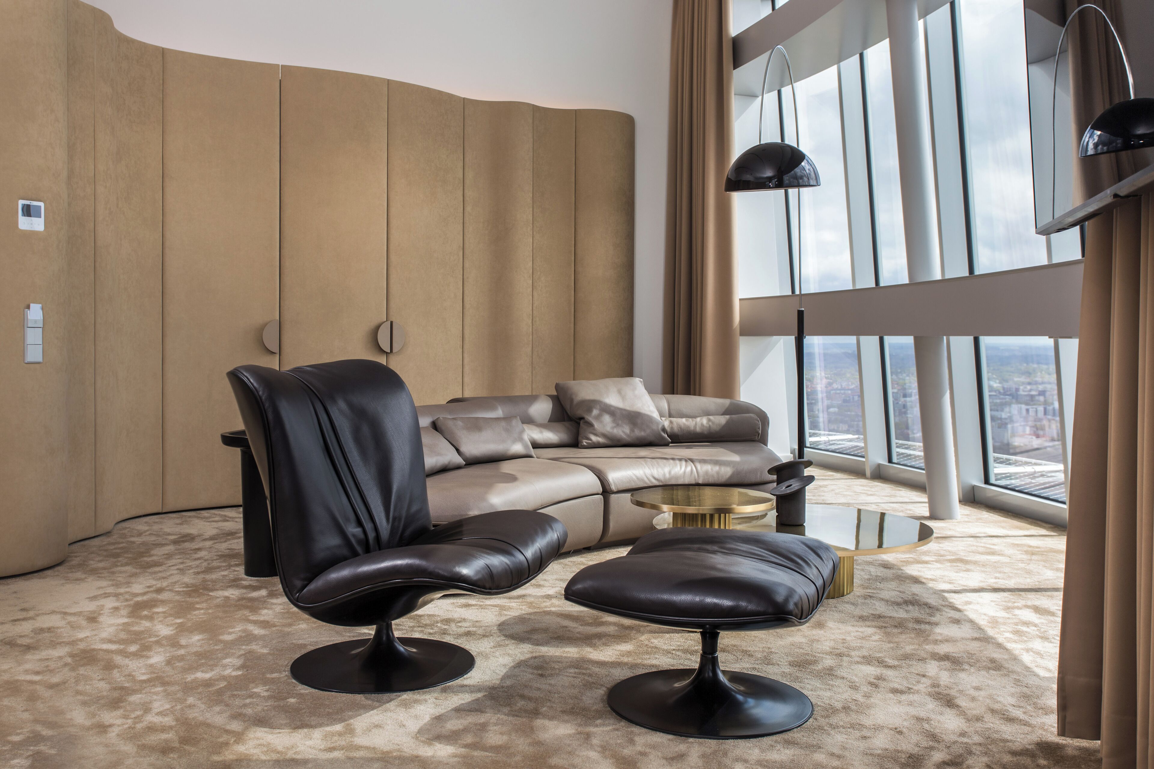 A modern living room with a leather sofa, two leather armchairs, a round coffee table, and a large window offering a city view.