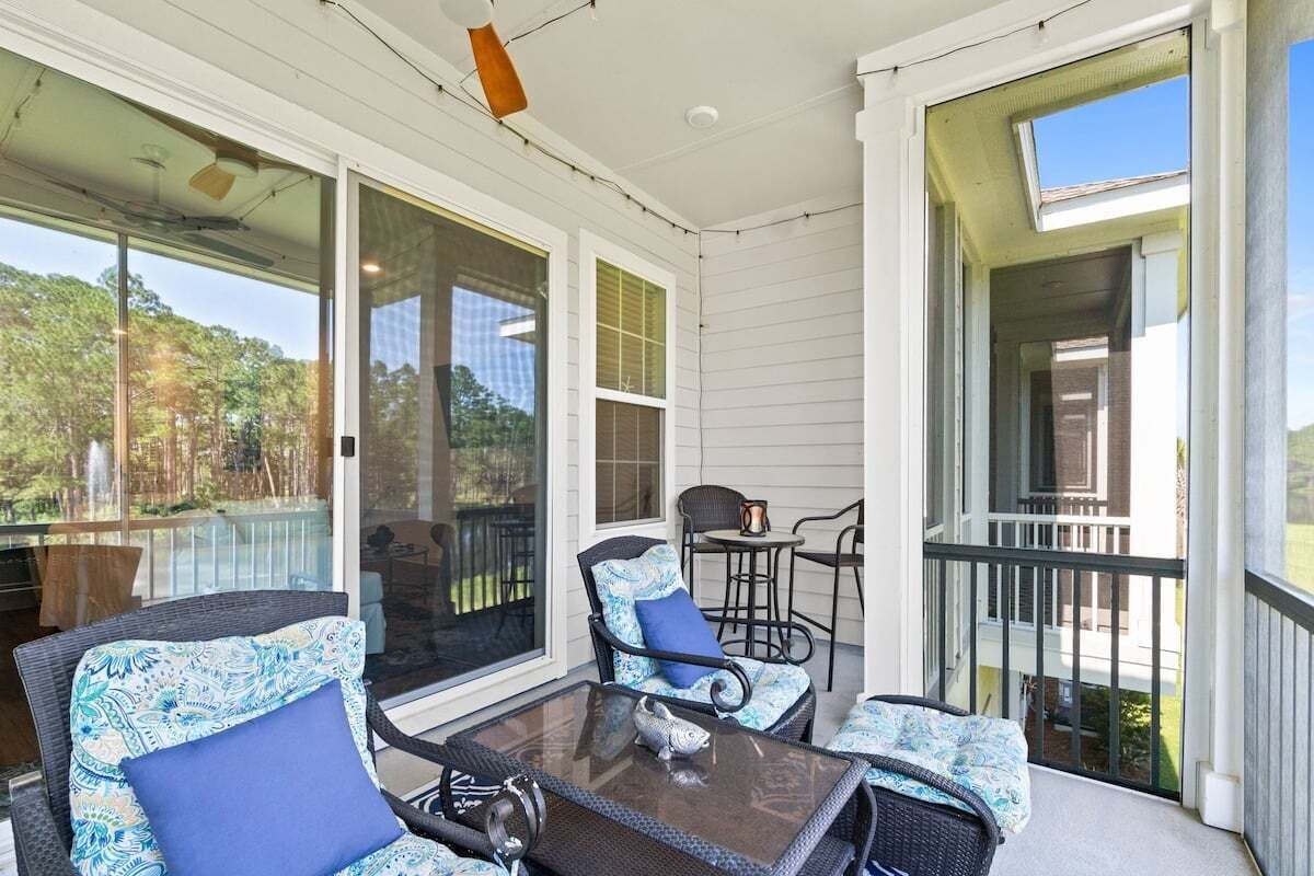 A covered porch with wicker furniture and a glass table, large windows, and a view of trees.