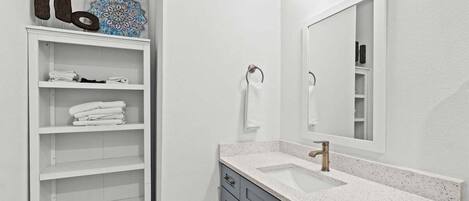 Un baño con tocador blanco, un espejo grande y estante con toallas dobladas.