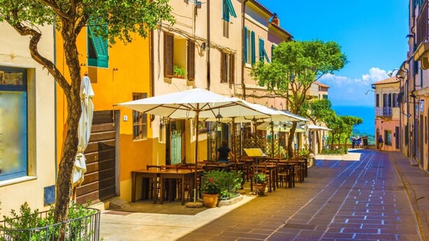 Una calle estrecha bordeada de cafés con terraza, con mesas y sillas bajo sombrillas, y edificios con balcones y contraventanas.