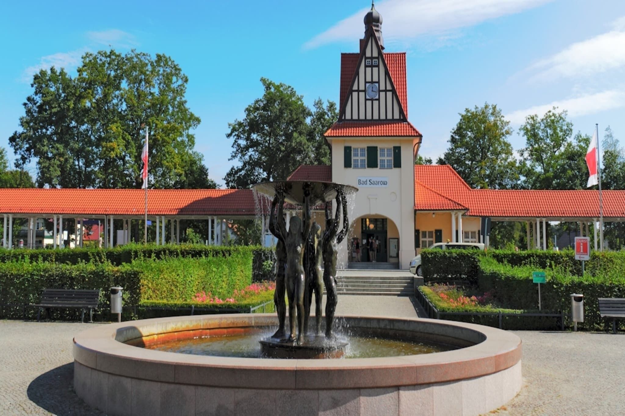 A public square with a fountain, a building with a clock tower, and a red-roofed structure.