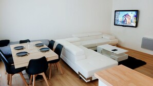 A modern living room with a dining area, white furniture, and a flat-screen TV displaying a cityscape. - Appartement T2 55m2 Plein Centre St-brieuc (Saint-Brieuc)