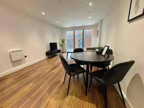 Quarto moderno com uma mesa redonda, quatro cadeiras, uma TV, um vaso de planta e uma janela com persianas.