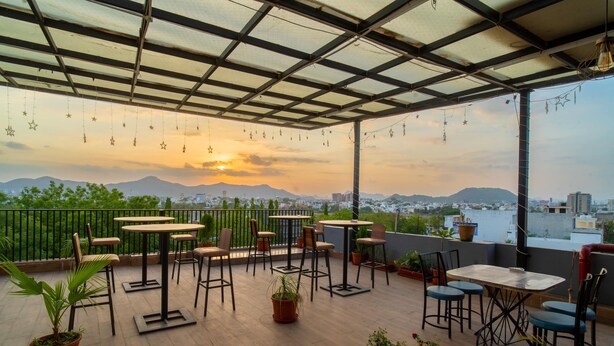 Une terrasse sur le toit, aménagée avec des tables et des chaises, des plantes en pot, et offrant une vue sur le paysage urbain et les montagnes au coucher du soleil.