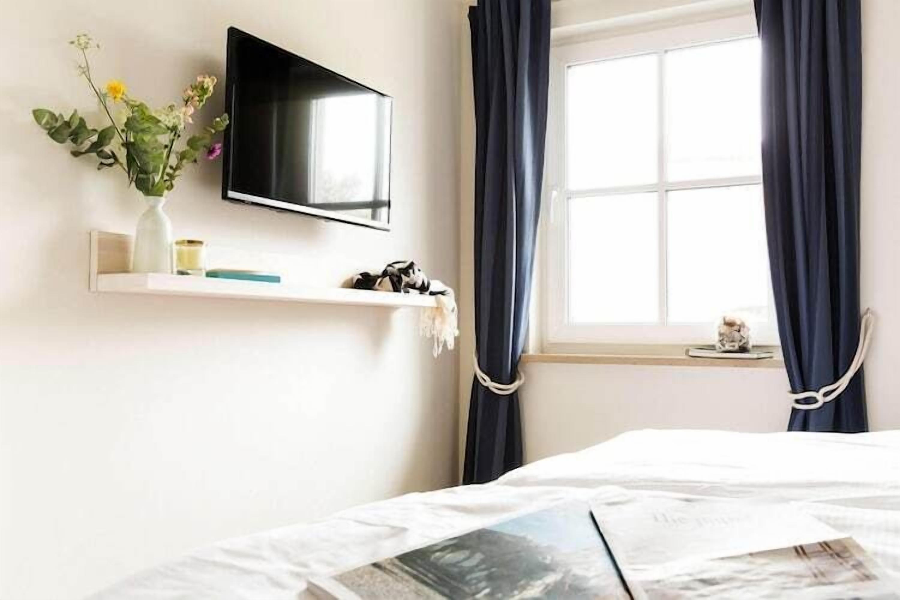 Une chambre à coucher avec un lit, un téléviseur fixé au mur, une fenêtre avec des rideaux et une petite étagère sur laquelle est posé un vase de fleurs.