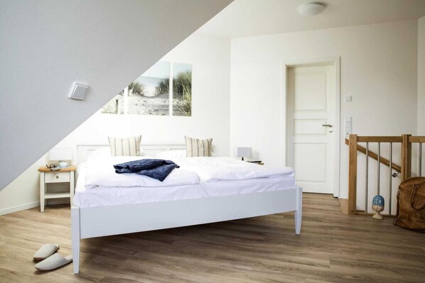 A bedroom with a white bed, a nightstand, a wooden floor, a door, and a view of the beach through a window. - Ferienwohnung, Zingst (Zingst)