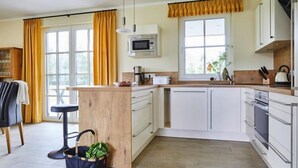 A modern kitchen with white cabinets, a wooden island, a microwave, and a window with yellow curtains. - Premium-Ferienhaus in Fuhlendorf (Fuhlendorf)