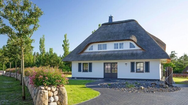 A house with a thatched roof, white exterior walls, and a stone wall with plants. - Premium-Ferienhaus in Fuhlendorf (Fuhlendorf)