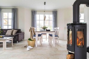 A living room with a sofa, a dining table with chairs, a fireplace, and large windows with curtains. - Haus am Bodden mit Sauna (Fuhlendorf)