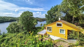A yellow cabin with a small deck and a solar panel, surrounded by lush greenery and a lake.