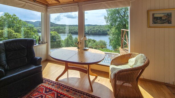 A sunlit room with a view of a river and forest, a round wooden table, a wicker chair, and a leather sofa.
