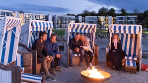 Un grupo de personas sentadas en sillas de playa a rayas alrededor de un brasero, con edificios modernos al fondo.
