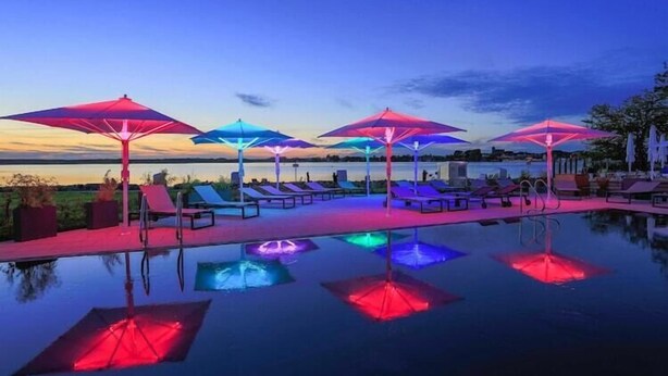 Zona de piscina con sillones reclinables y sombrillas, con reflejos en el agua.