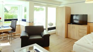 Une chambre d’hôtel moderne équipée d’un canapé, d’un téléviseur posé sur un meuble en bois, d’un coin repas avec une table et des chaises, et d’une grande fenêtre donnant sur un paysage verdoyant.
