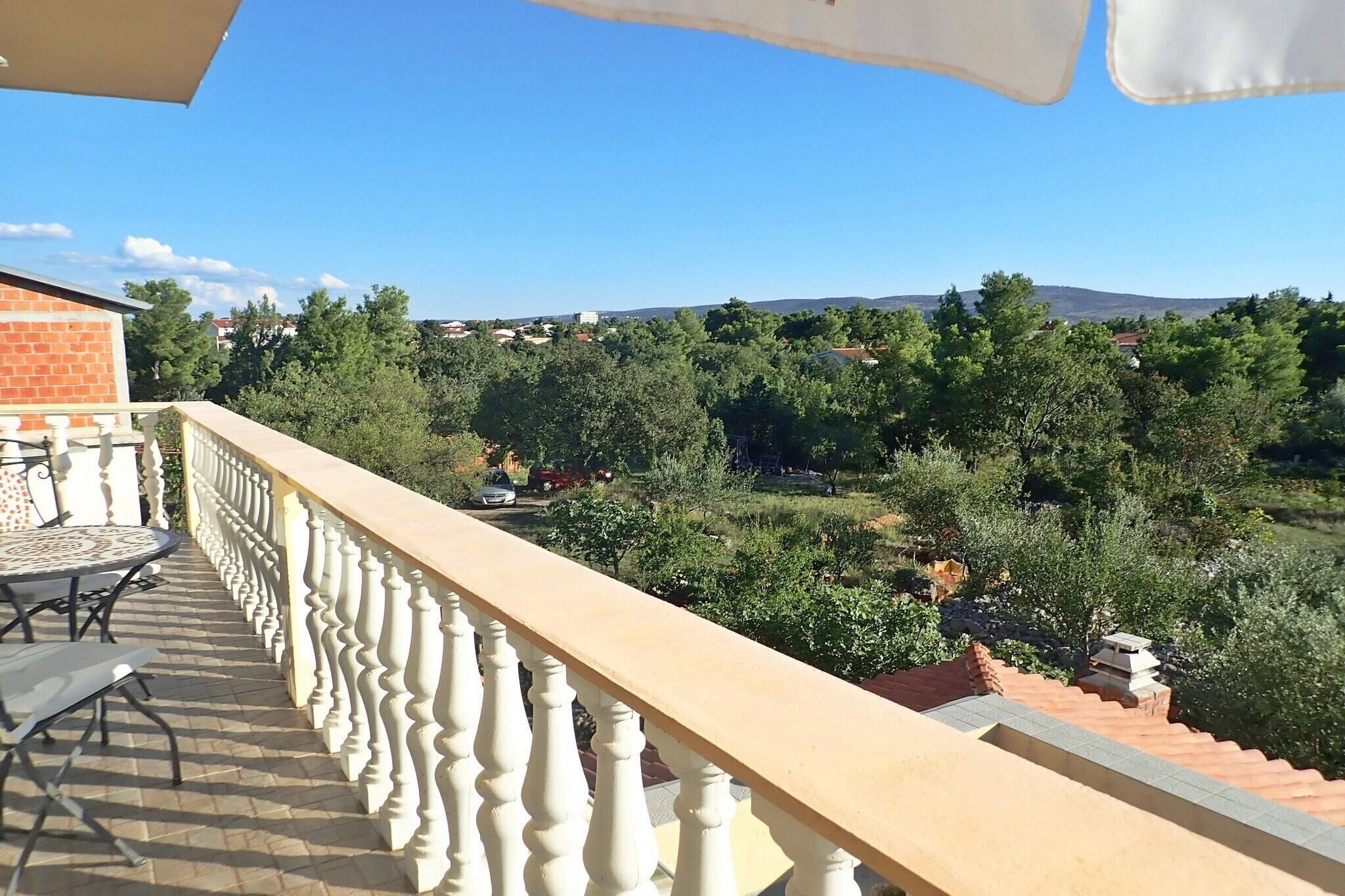 Ein Balkon mit weißer Brüstung, einem Tisch mit Stühlen und Blick auf eine grüne Landschaft mit Bäumen und Häusern.