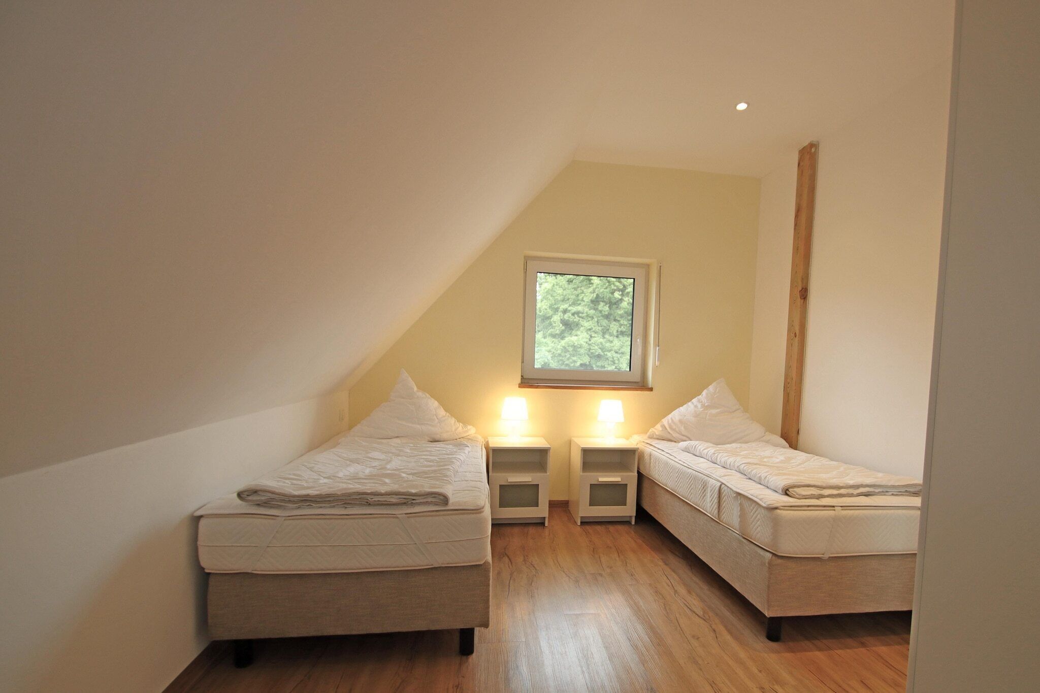 A small, single-room with two beds, a window, and a sloped ceiling.