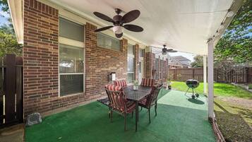 A covered patio with a brick wall, a ceiling fan, a table with chairs, and a grill.