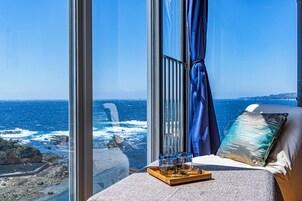 A room with a large window overlooking the ocean, a bed with pillows, and a tray with glasses.