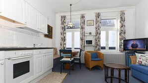 Una cocina funcional con armarios blancos, horno integrado y comedor con mesa y sillas. En la zona de estar hay un televisor, una silla de mimbre y una pequeña mesa redonda.