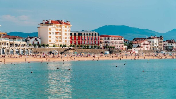 Un pueblo costero con una playa, edificios y montañas al fondo.