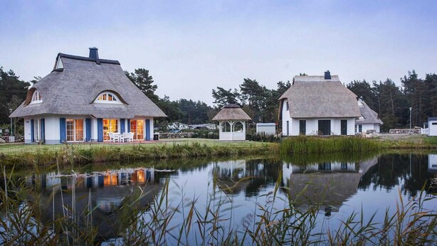 A row of thatched-roof cottages with a calm pond in the foreground. - Ferienhaus in Fuhlendorf (Fuhlendorf)