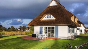A thatched-roof house with a white exterior, a patio with outdoor furniture, and a pond in the background. - Ferienhaus in Fuhlendorf (Fuhlendorf)
