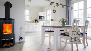 A cozy living room with a wood-burning stove, a dining area with a table and chairs, and a kitchen in the background. - Ferienhaus, Fuhlendorf (Fuhlendorf)