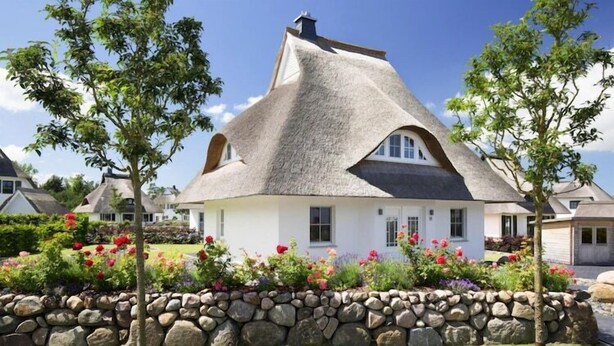 A house with a unique thatched roof, surrounded by a stone wall and flowering plants. - Ferienhaus, Fuhlendorf (Fuhlendorf)