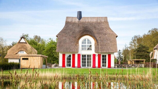 A house with a thatched roof and red shutters, surrounded by a pond and greenery.
