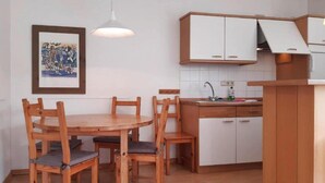 Een kleine keuken met witte kastjes, een houten eettafel met stoelen en een hangende hanglamp.