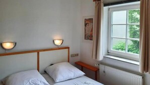 Een kamer met twee eenpersoonsbedden, een houten hoofdbord, wandlampen, een raam met gordijnen en een radiator.