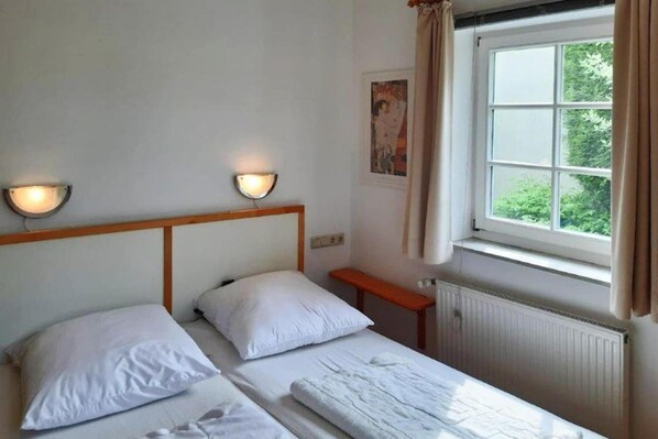 Habitación con dos camas individuales, cabecero de madera, luces fijadas a la pared, ventana con cortinas y un radiador.