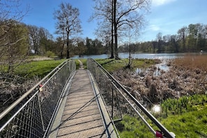 Un puente de madera con barandillas metálicas que conduce a una masa de agua, rodeado de árboles y zonas verdes.