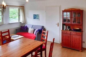 Ein Zimmer mit einem Holztisch, roten Stühlen, einem roten Sofa, einem Holzkasten und einem Fenster mit Vorhängen.