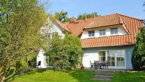 Une maison à deux étages, avec un toit recouvert de tuiles rouges, une grande véranda à l’avant dotée de grandes fenêtres et une pelouse bien entretenue.